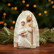 Decorative nativity scene wood carving with Joseph holding a staff next to Virgin Mary holding Baby Jesus on a table with festive Christmas background