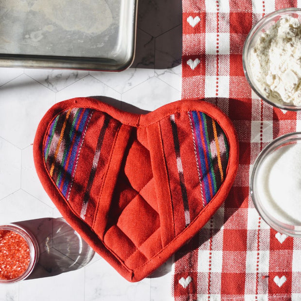 Heart-shaped festive red pot holder next to a checkered tablecloth with ingredients around