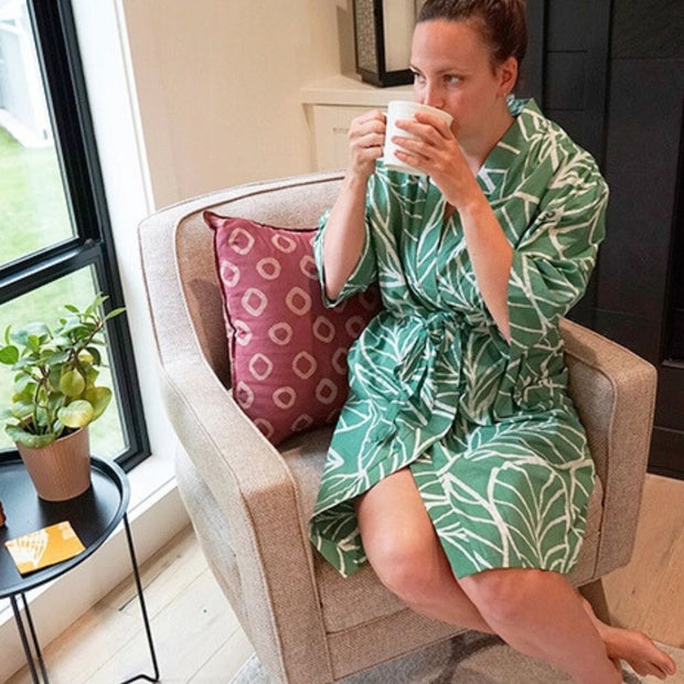 Woman in a green patterned robe from Global Mamas sitting on a chair holding a mug in a cozy living room.